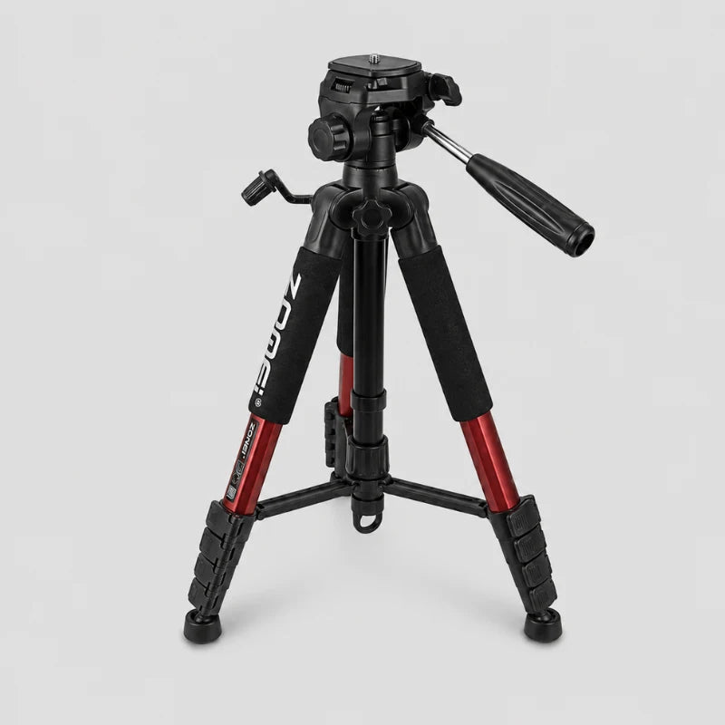 Trépied Zomei rouge et noir en position très basse sur fond gris, tête panoramique 3 voies noire avec levier, grips mousse noirs, jambes rouges écartées, vue trois quarts